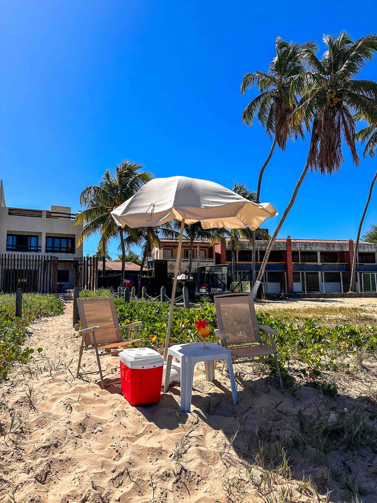 Two chairs sit under a white umbrella on a sandy beach, accompanied by a red cooler. Lush green foliage is visible surrounding the area, while swaying palm trees tower in the background. The clear blue sky creates a bright backdrop to the beach setting.