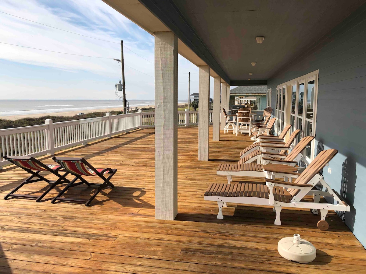 An expansive wooden deck offers direct views of the beach, furnished with multiple lounge chairs and a couple of deck chairs. Sunlight streams across the surface, highlighting the space available for relaxation and enjoyment of the coastal scenery.