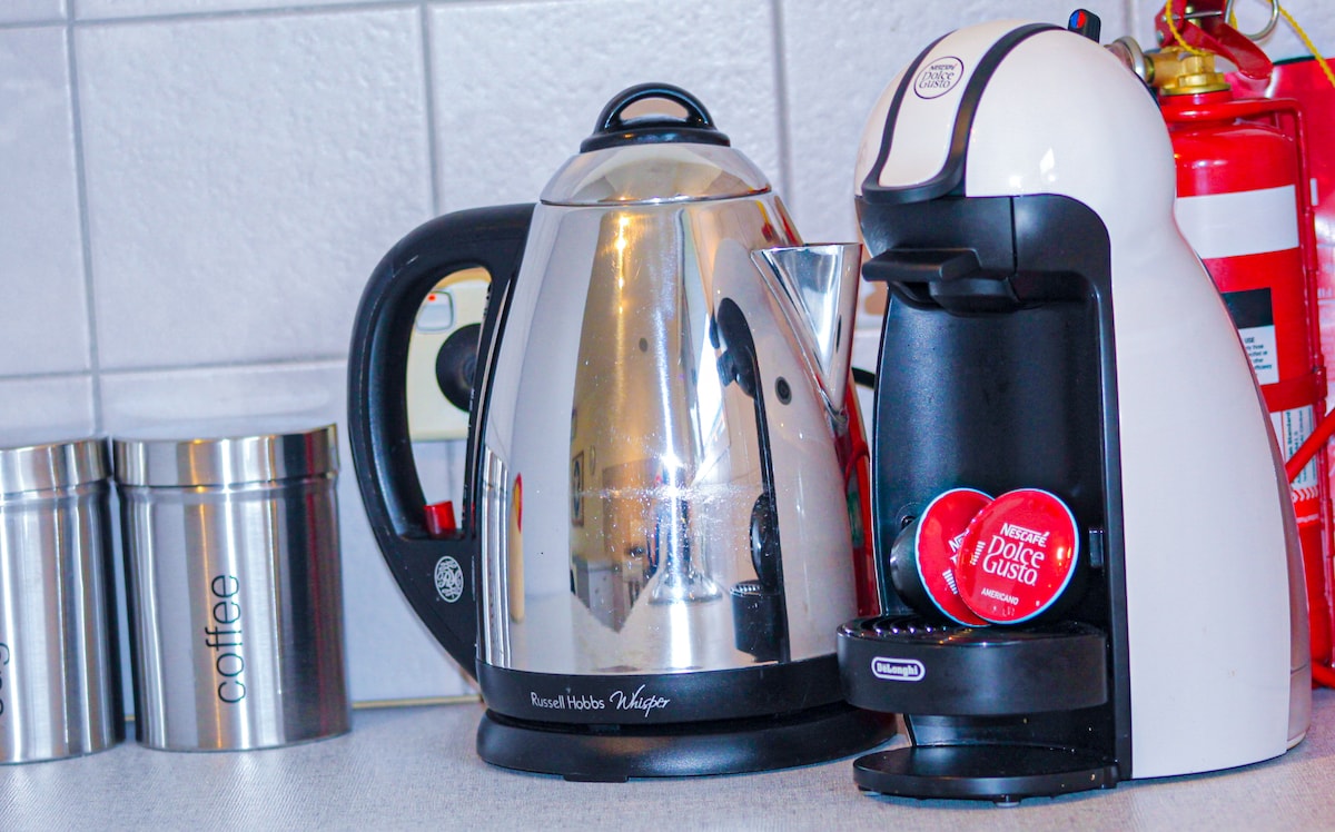A stainless steel kettle is positioned next to a coffee machine, featuring a sleek design. Two coffee capsules are visible, emphasizing the availability of coffee-making amenities. The countertop is clear, adding to the organized appearance of the kitchen space.
