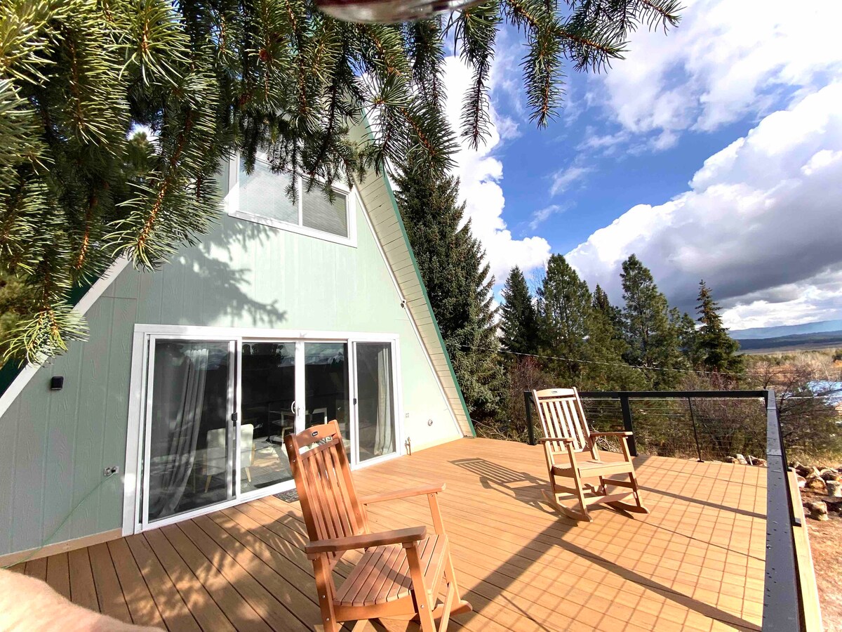 The outdoor deck of the cabin is shown, featuring two wooden rocking chairs positioned for enjoying views of the surrounding trees and mountains. Natural light filters through the green A-frame structure, creating a welcoming outdoor space.