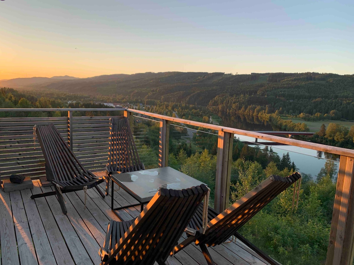 A spacious deck features two lounge chairs positioned for optimal views of the river and surrounding landscape. A low coffee table rests between the chairs, enhancing the serene setting during sunset. The robust wooden railings frame the picturesque outdoor space.