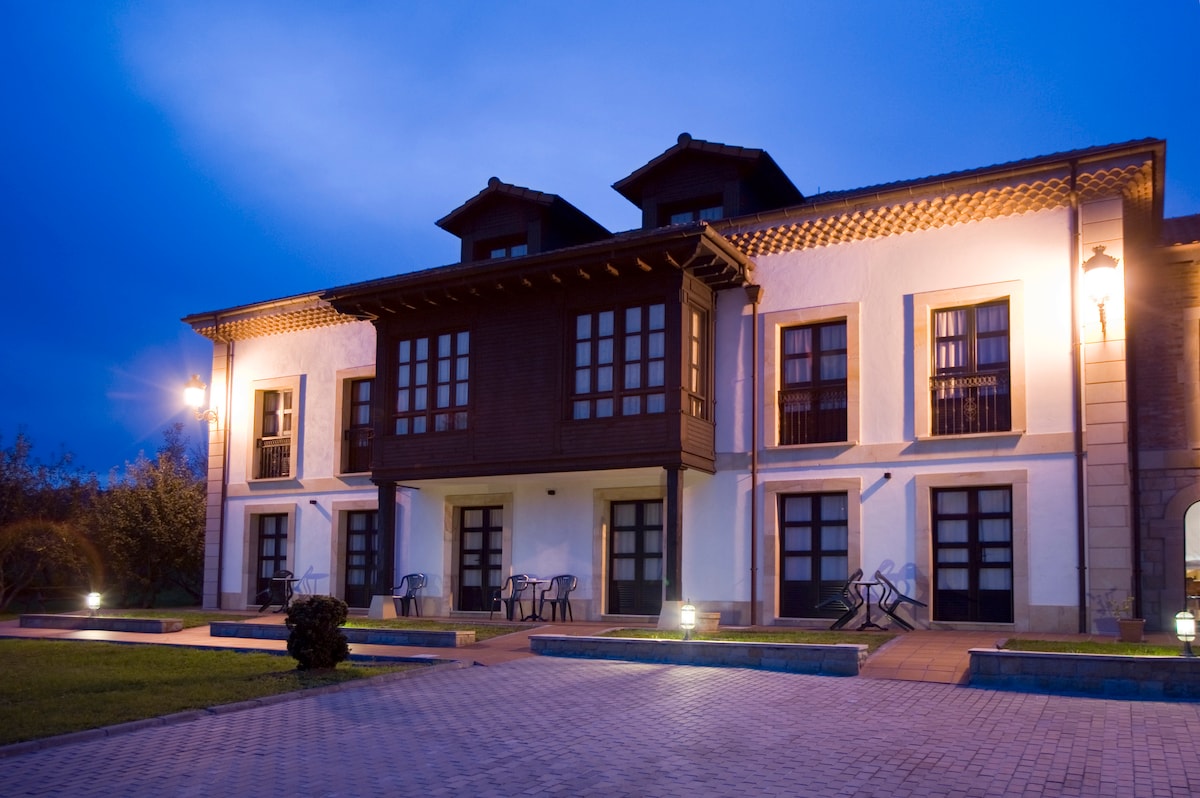 An elegant building is depicted at dusk, featuring a wooden balcony and ground-floor terraces with seating. Soft outdoor lighting illuminates the stone pathway and landscaped areas, while the serene sky adds a tranquil backdrop to the architectural design.