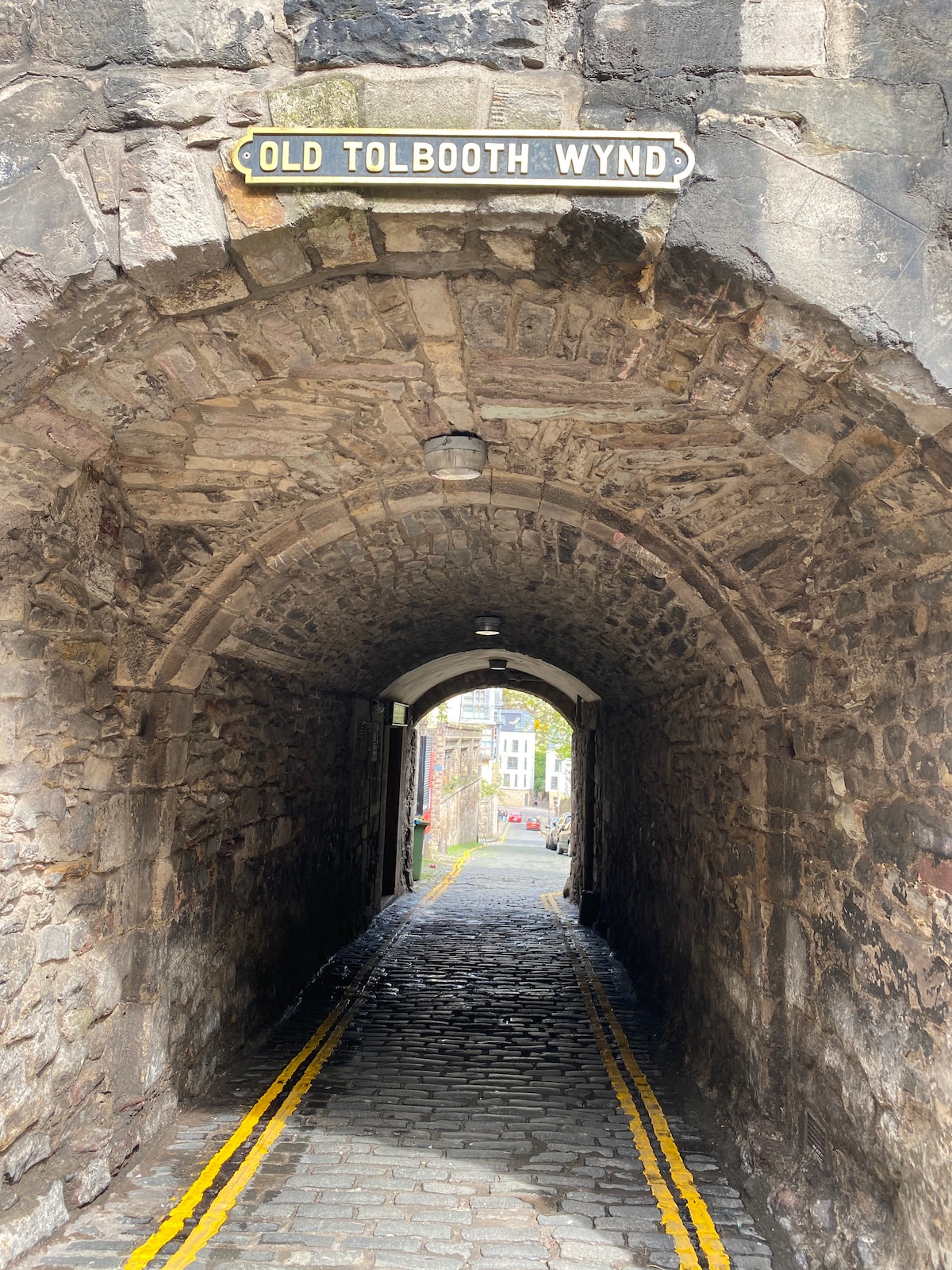 A stone archway leads towards a cobblestone path marked by double yellow lines. The sign overhead indicates 'OLD TOLBOOTH WYND.' Natural light filters through the arch, illuminating the rugged stone walls, which exhibit historical character and charm.
