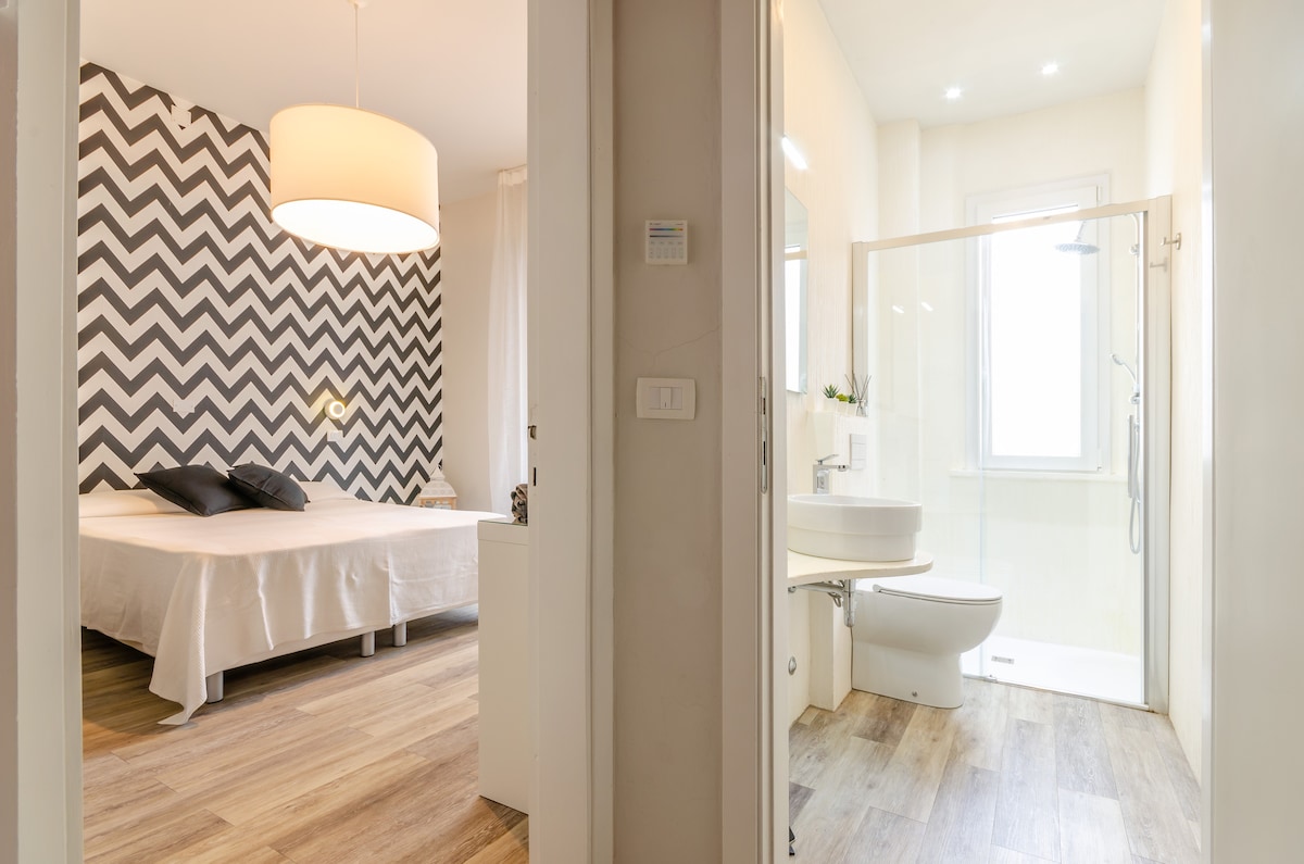 A bright bedroom features a spacious bed with black accent pillows against a patterned chevron wall. An adjoining bathroom is visible, showcasing a modern shower, sink, and toilet. Natural light filters through a window, enhancing the airy feel of the space.