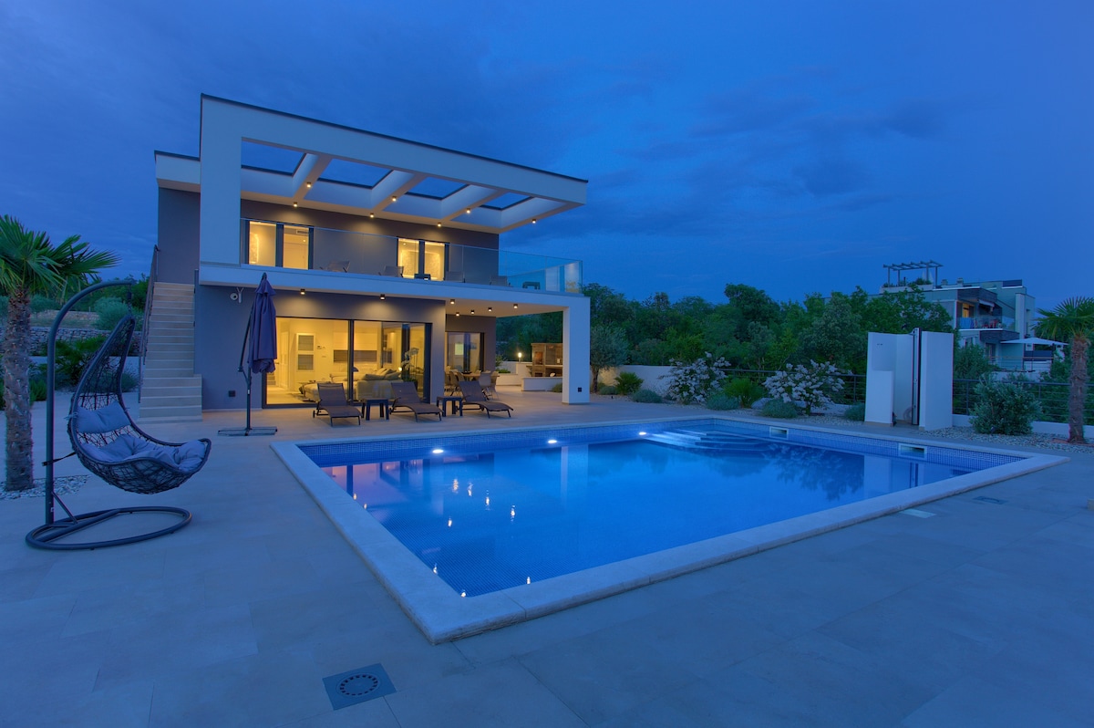 The modern villa is presented at dusk, showcasing a private heated swimming pool illuminated by soft lights. Comfortable outdoor seating is arranged nearby under a shaded area, while lush greenery enhances the tranquil environment.