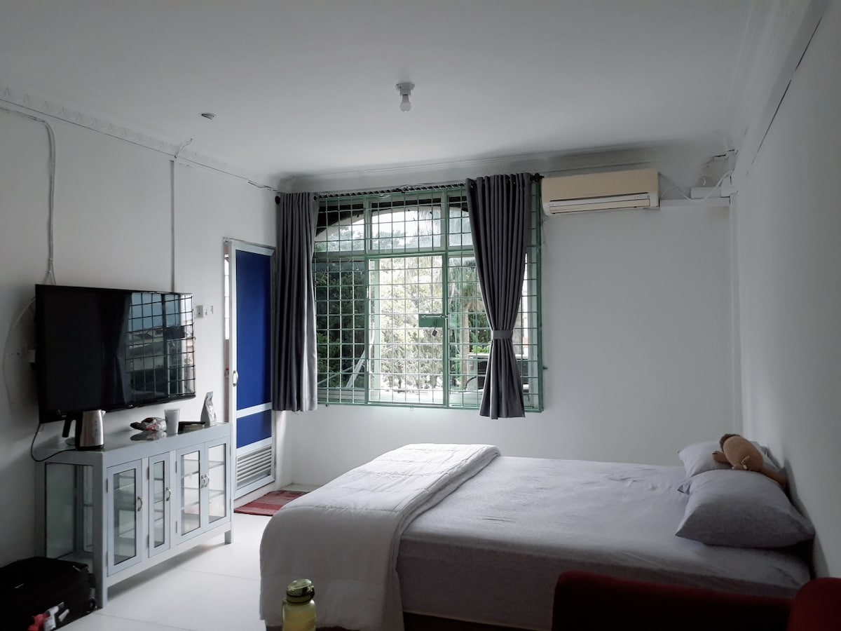 A spacious room features a large bed with neutral bedding and a small area with a plush chair. A flat-screen TV is mounted on the wall opposite the bed. Natural light enters through a grid-patterned window, complemented by light curtains, creating a bright and airy atmosphere.