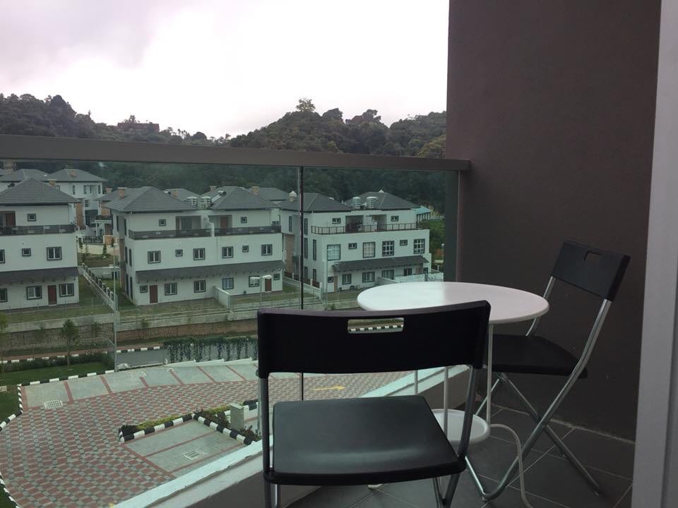 A balcony is shown, featuring a small round table and two black chairs. The view includes neighboring buildings and greenery, with a cloudy sky in the background. The modern glass railing adds to the open feel of the outdoor space.