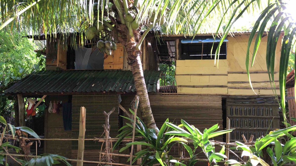 A rustic wooden cabin stands surrounded by lush greenery, featuring two levels beneath a thatched roof. Openings on the upper level allow for natural light, while the lower area exhibits bamboo siding. Palm trees frame the space, enhancing the tropical ambiance.