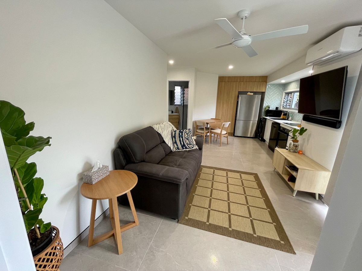 An open living area features a dark sofa paired with a small round table and four chairs. A large TV is mounted on the wall, and a cooling fan is visible on the ceiling. Natural light brightens the space, complemented by a textured area rug on the tiled floor.