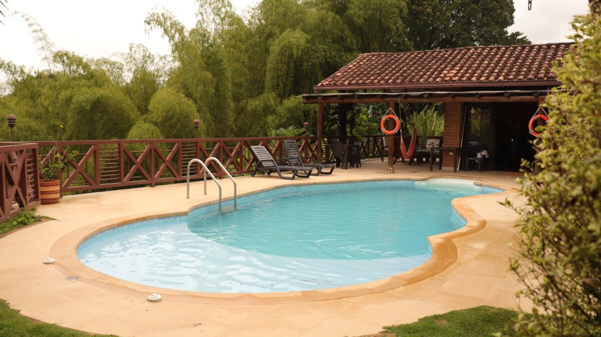 An inviting private pool is surrounded by landscaped greenery, featuring a spacious patio area with lounge chairs. A covered gazebo is visible in the background, furnished for outdoor dining and relaxation. The tranquil setting is enhanced by natural foliage and a clear sky.