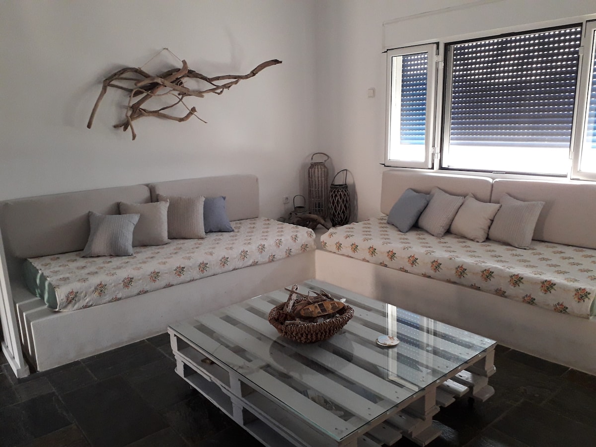 A spacious living area is furnished with two corner sofas featuring floral-patterned cushions. A glass coffee table sits centrally, adorned with a decorative bowl. Natural light filters through the large windows, enhancing the minimalist aesthetic of the room.