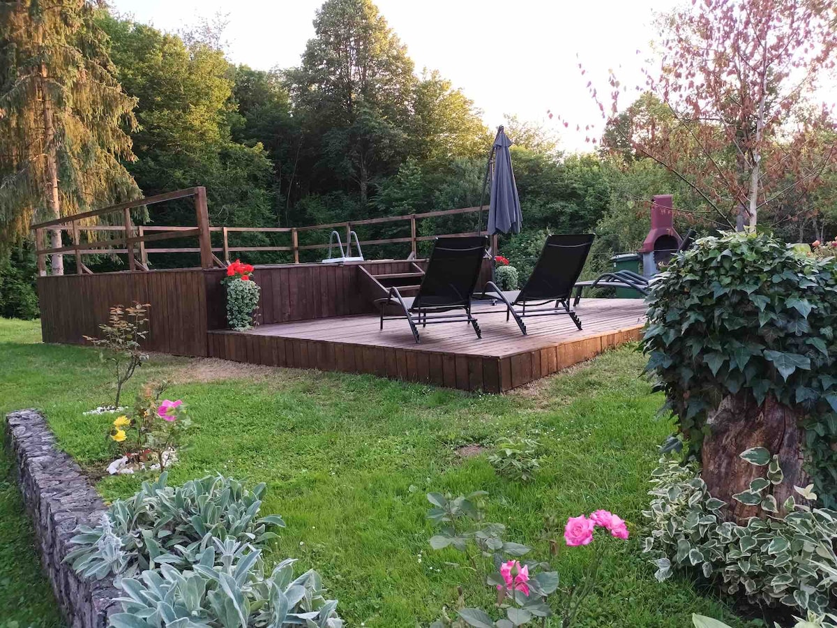 An outdoor deck features two lounge chairs positioned under a shaded umbrella. Lush greenery surrounds the space, with flower beds showcasing pink and white blooms. A wooden railing encloses the area, creating a serene environment for relaxation.