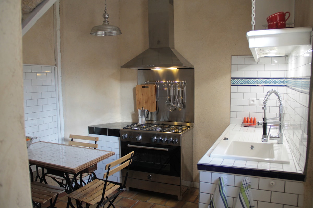 A modern kitchen is presented with a gas stove, oven, and stainless steel hood. A white sink is visible alongside a tiled backsplash. A wooden dining table with two folding chairs occupies one side, and kitchen utensils are neatly organized on the wall.