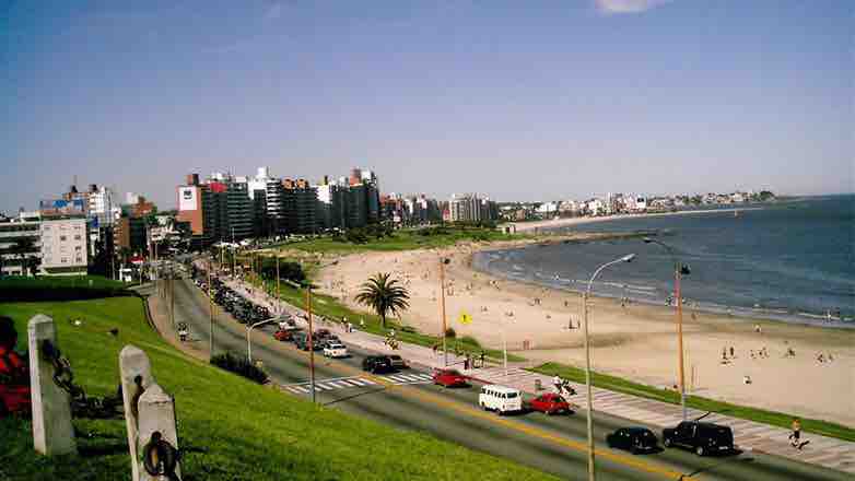 A scenic view captures a bustling beach area bordered by tall buildings. A sandy shoreline meets gentle waves, while people are seen enjoying leisure activities. A road lined with vehicles runs parallel to the beach, adding to the lively atmosphere of this coastal location.