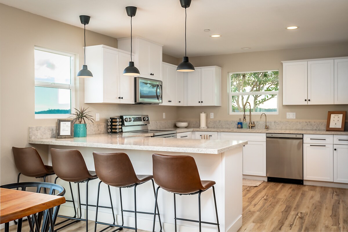 The kitchen features modern white cabinetry and granite countertops, complemented by four brown bar stools at the island. Stainless steel appliances, including a microwave and dishwasher, are visible. Natural light enters through the two windows, illuminating the space.