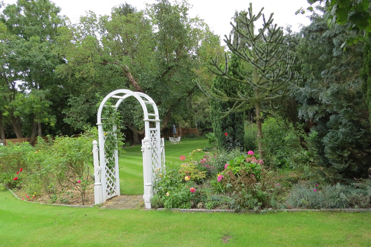 A charming garden area is showcased with a white wooden archway leading into a lush green space. Colorful flowers bloom along the pathways, and well-maintained hedges create a serene atmosphere. A seating area with white chairs can be seen in the distance, surrounded by trees.