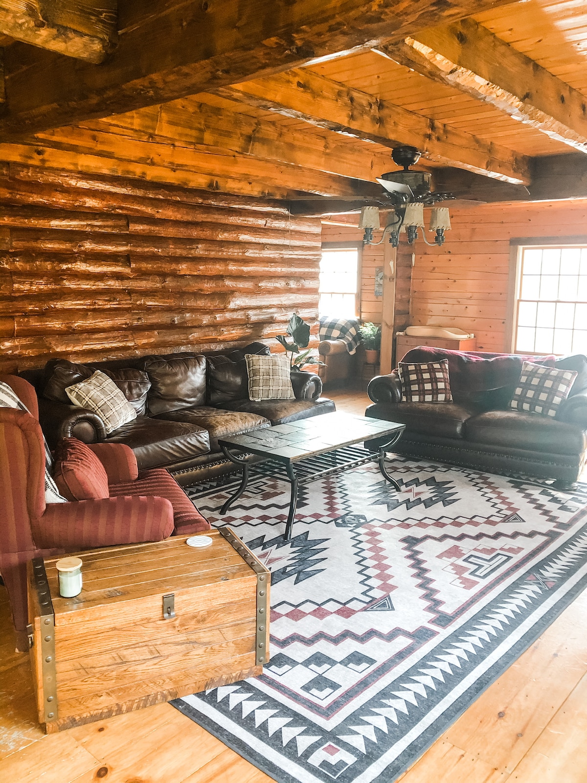 A cozy living area features rich log walls and wooden beams overhead. Comfortable seating includes a leather couch and armchairs, arranged around a central table on a patterned rug. Natural light floods the space through large windows, enhancing the inviting atmosphere.