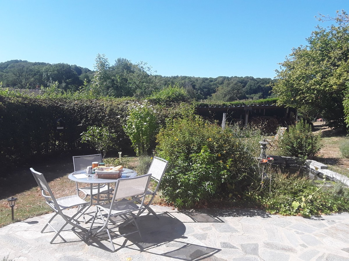 A patio area is presented with a circular table surrounded by four chairs, set against a backdrop of greenery. A tranquil garden scene is highlighted by flowering plants and shrubbery, with sunlight casting a warm glow on the stone surface.