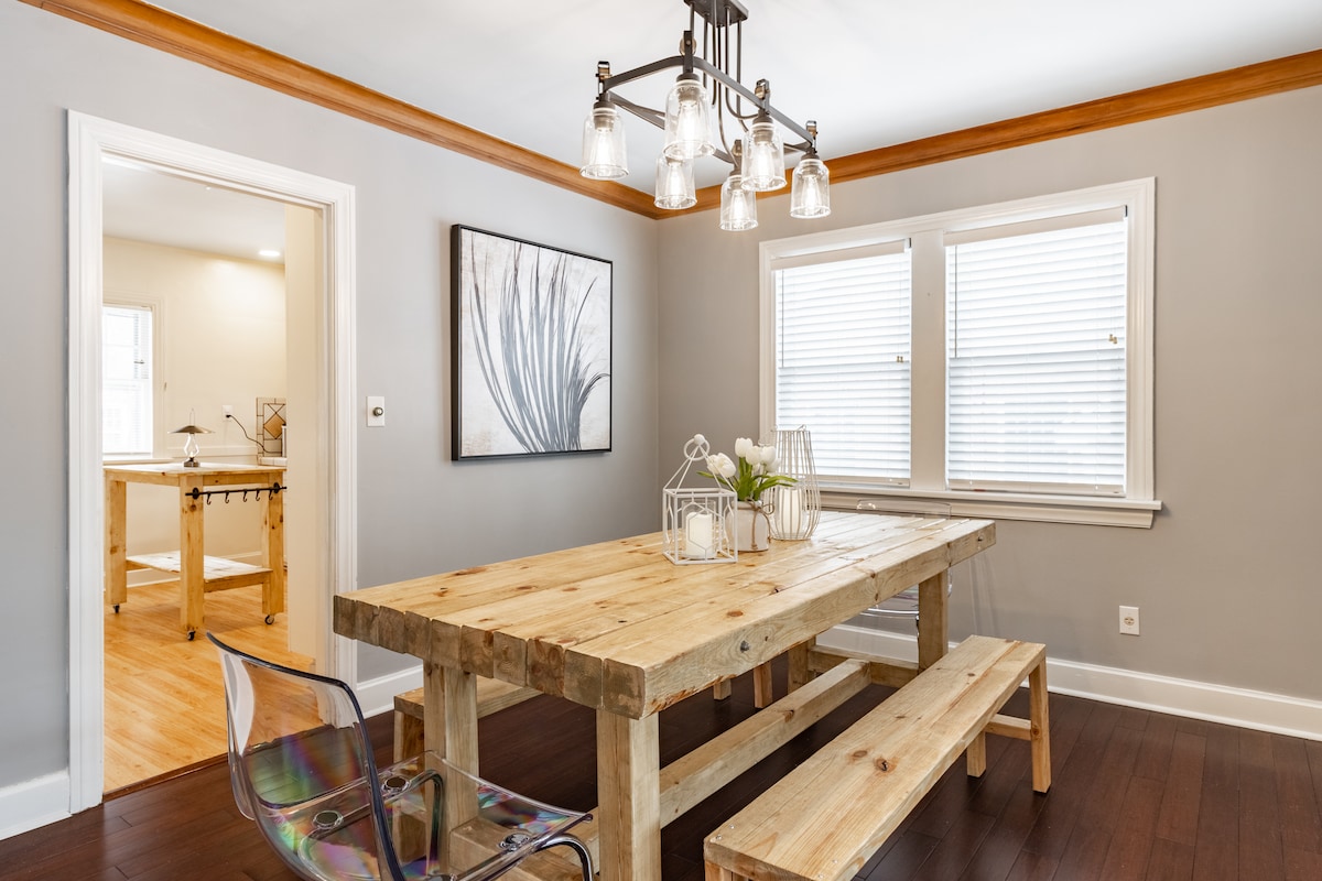 A dining area features a large wooden table surrounded by a bench and clear acrylic chairs. Natural light streams through two windows adorned with white blinds. A modern light fixture hangs overhead, complemented by a piece of wall art showcasing botanical elements.