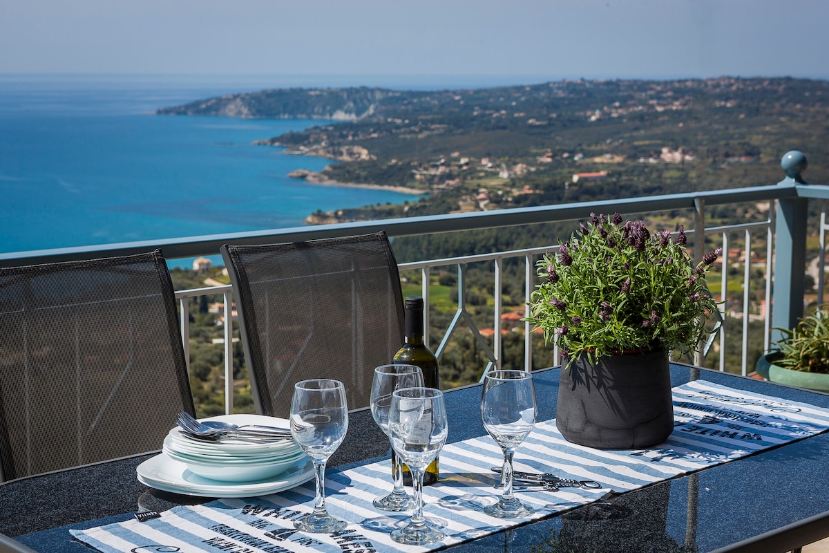 A dining table set for four features neatly arranged plates and glasses, placed on a patterned table runner. A potted plant adds a touch of greenery, while a bottle sits nearby. The background offers views of the sea and surrounding landscape.