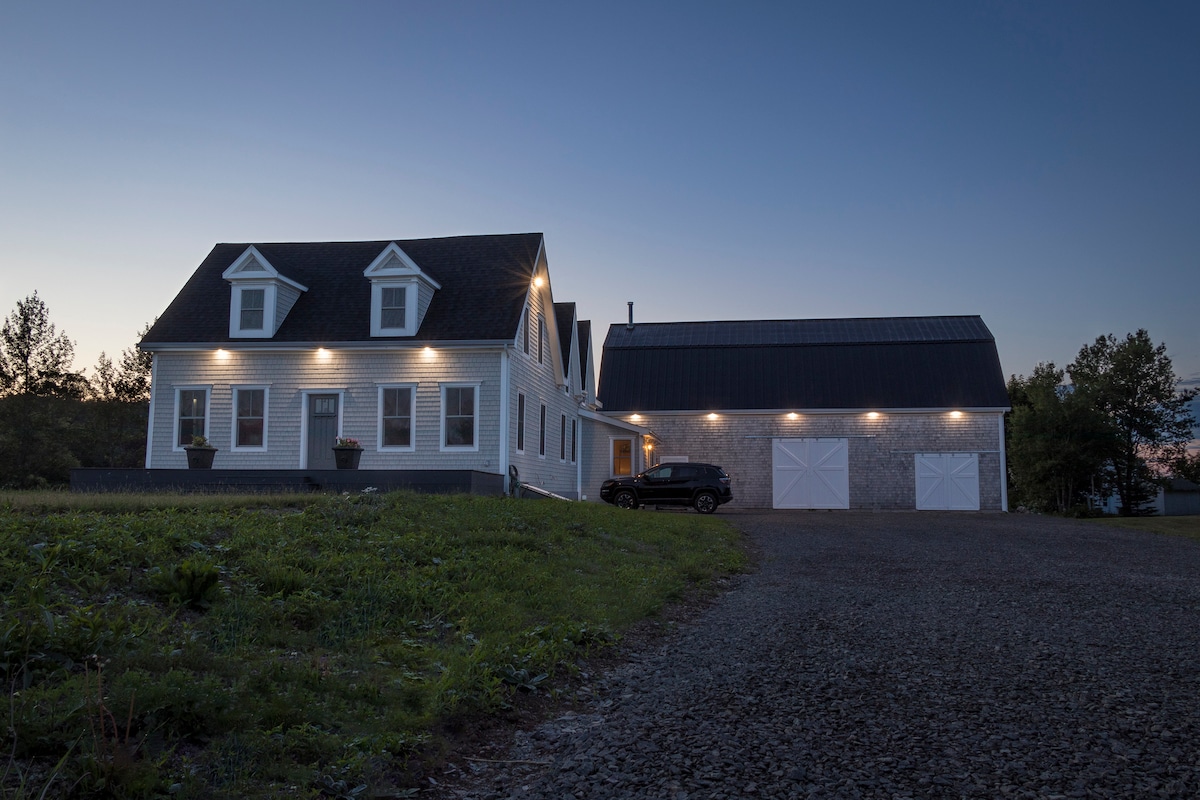 The newly renovated farmhouse is shown at dusk, featuring a traditional architecture with a grey shingle roof and bright siding. Warm lights illuminate the exterior, highlighting the inviting entrance and landscaping. A spacious gravel driveway leads to a parking area beside the house and attached building.