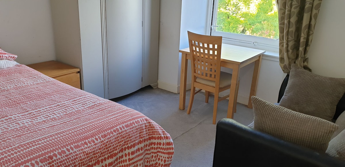 A comfortable bedroom includes a spacious bed with a patterned bedspread. Natural light filters through a nearby window, illuminating a wooden table with a grid-patterned chair. A soft armchair is positioned next to the window, enhancing the cozy ambiance.