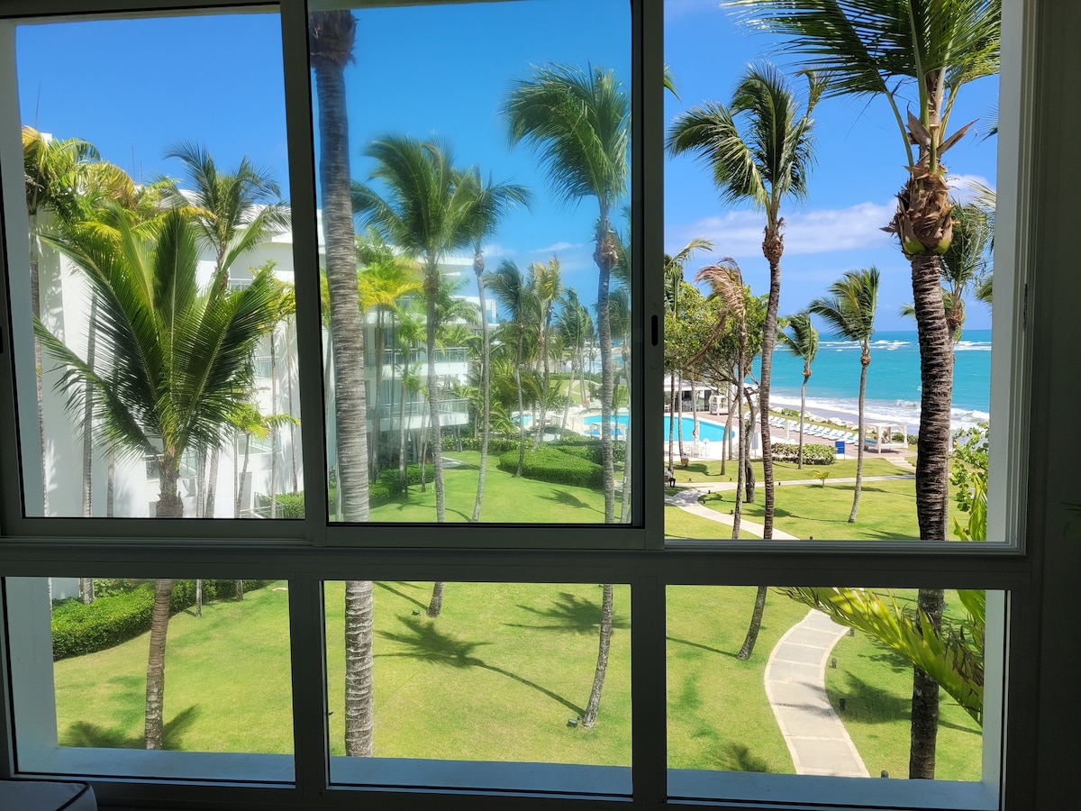 A bright view through large glass windows showcases lush palm trees swaying gently in the breeze. A turquoise pool is visible in the background, flanked by well-maintained green areas. The ocean glistens just beyond the pool, creating a serene coastal atmosphere.