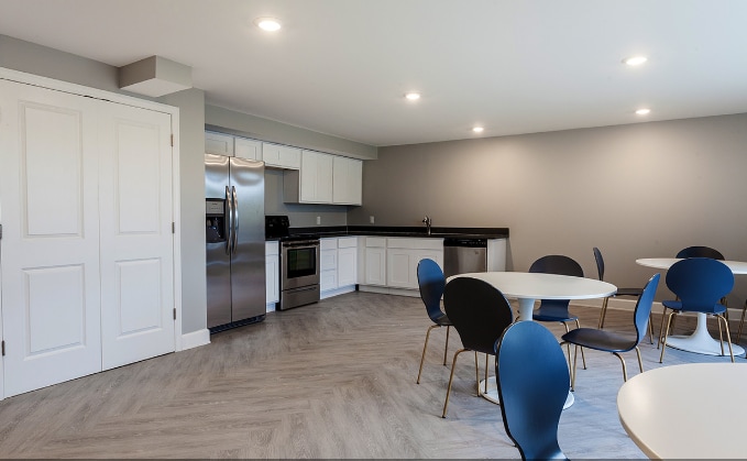 A spacious kitchen area showcases modern appliances, including a stainless steel refrigerator and oven. Dining tables with round tops and black and blue chairs are arranged to offer ample seating. Wood-like flooring enhances the light-filled environment, while cabinets provide storage space.