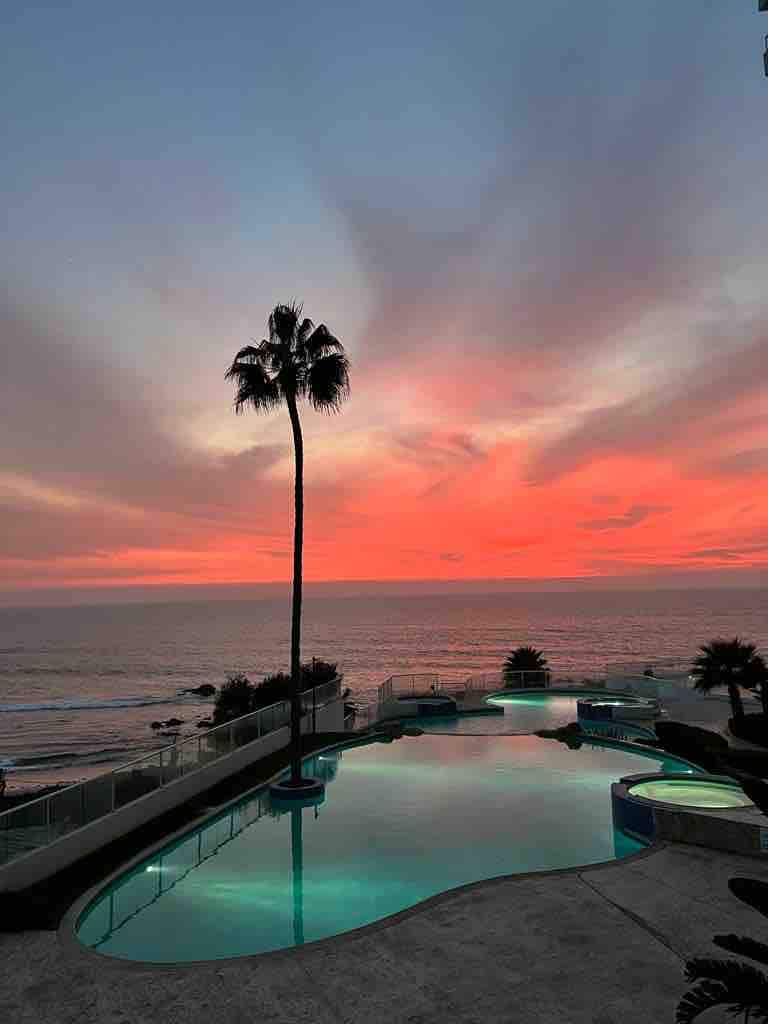 A serene view of the ocean at sunset is framed by a palm tree and a winding pool. The sky displays vibrant hues of orange and pink, reflecting on the water's surface. The peaceful atmosphere is complemented by gentle ocean waves visible in the distance.