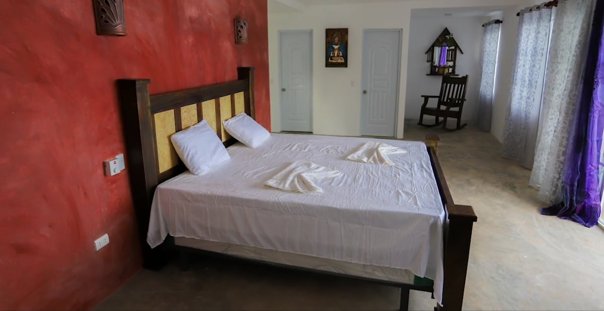 A spacious sleeping area features a large bed with white linens and two pillows neatly arranged. Soft red walls enhance the vibrant decor. Natural light enters through curtains framing a window, and a wooden rocking chair is visible in the corner of the room.