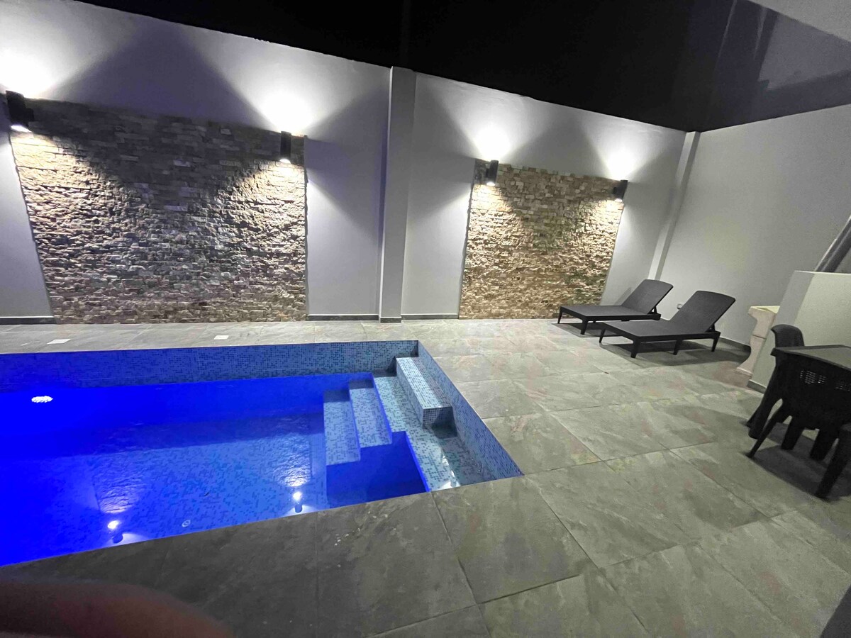 A private pool area features a striking blue water pool, accompanied by smooth stone walls illuminated by soft lighting. Two lounge chairs are positioned beside the pool, inviting relaxation in the tranquil surroundings.