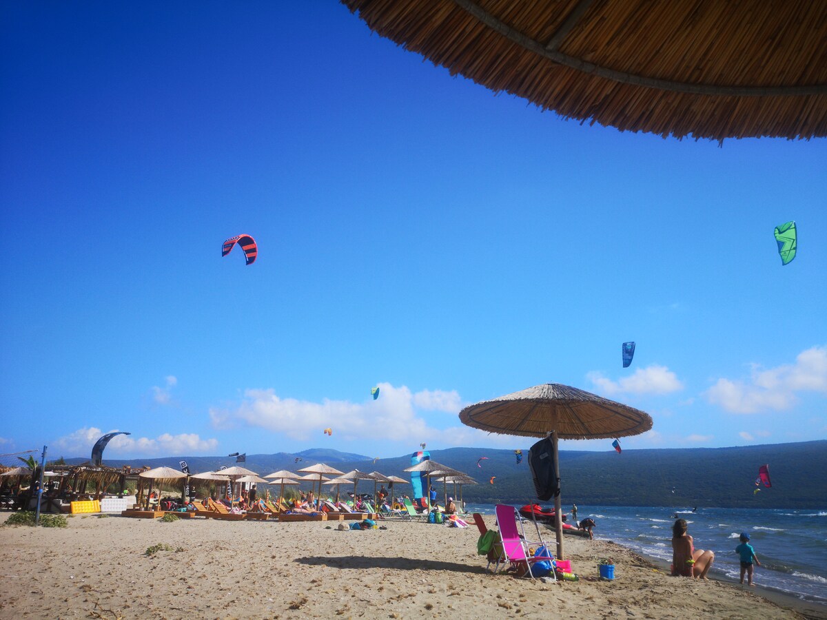 A sandy beach scene features a variety of vibrant kites soaring in the blue sky above. Rows of sun loungers and umbrellas provide shaded areas for relaxation. The shoreline gently meets the water, with distant hills creating a serene backdrop.