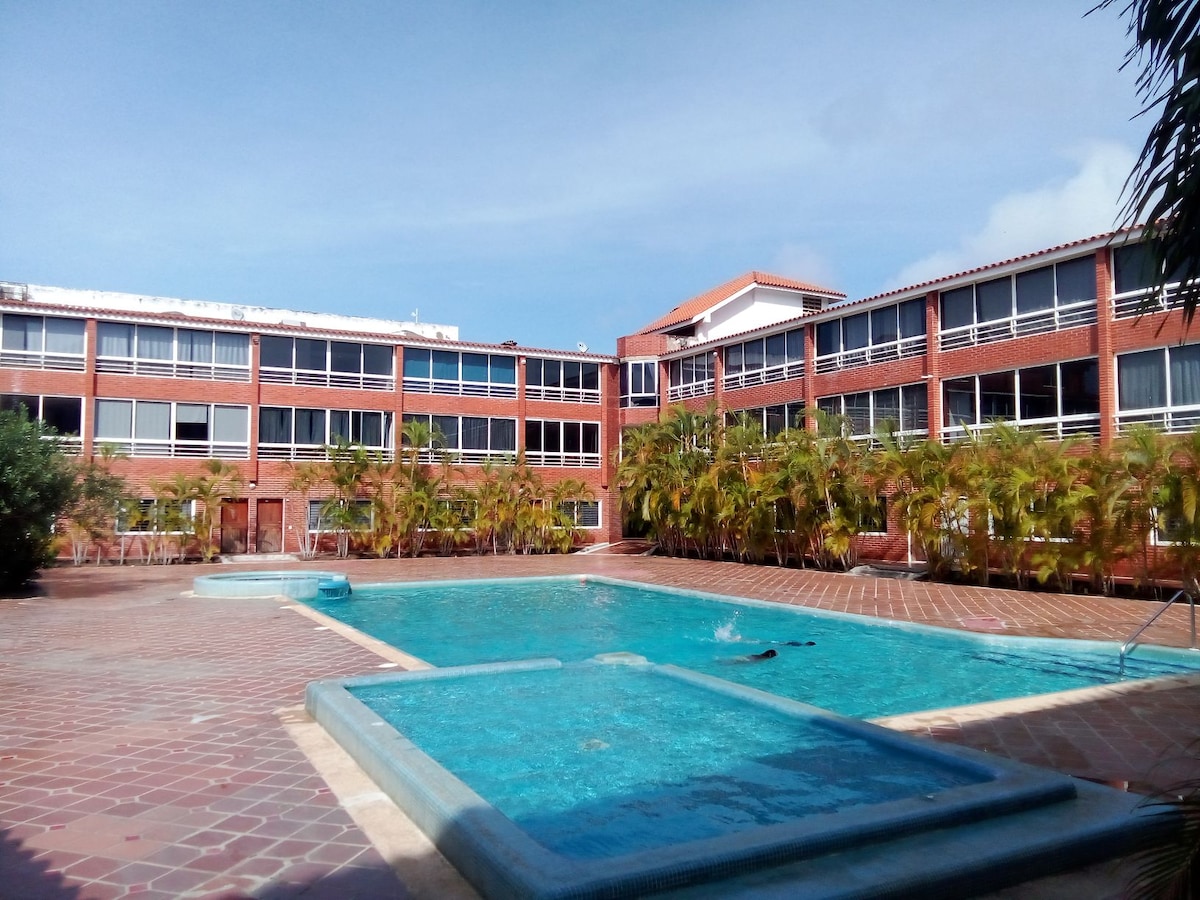 A serene outdoor area features a clear swimming pool surrounded by brick architecture. Lush greenery lines the perimeter, with spacious walkways leading around the pool. Bright windows are visible across the building, providing a sense of openness and light.