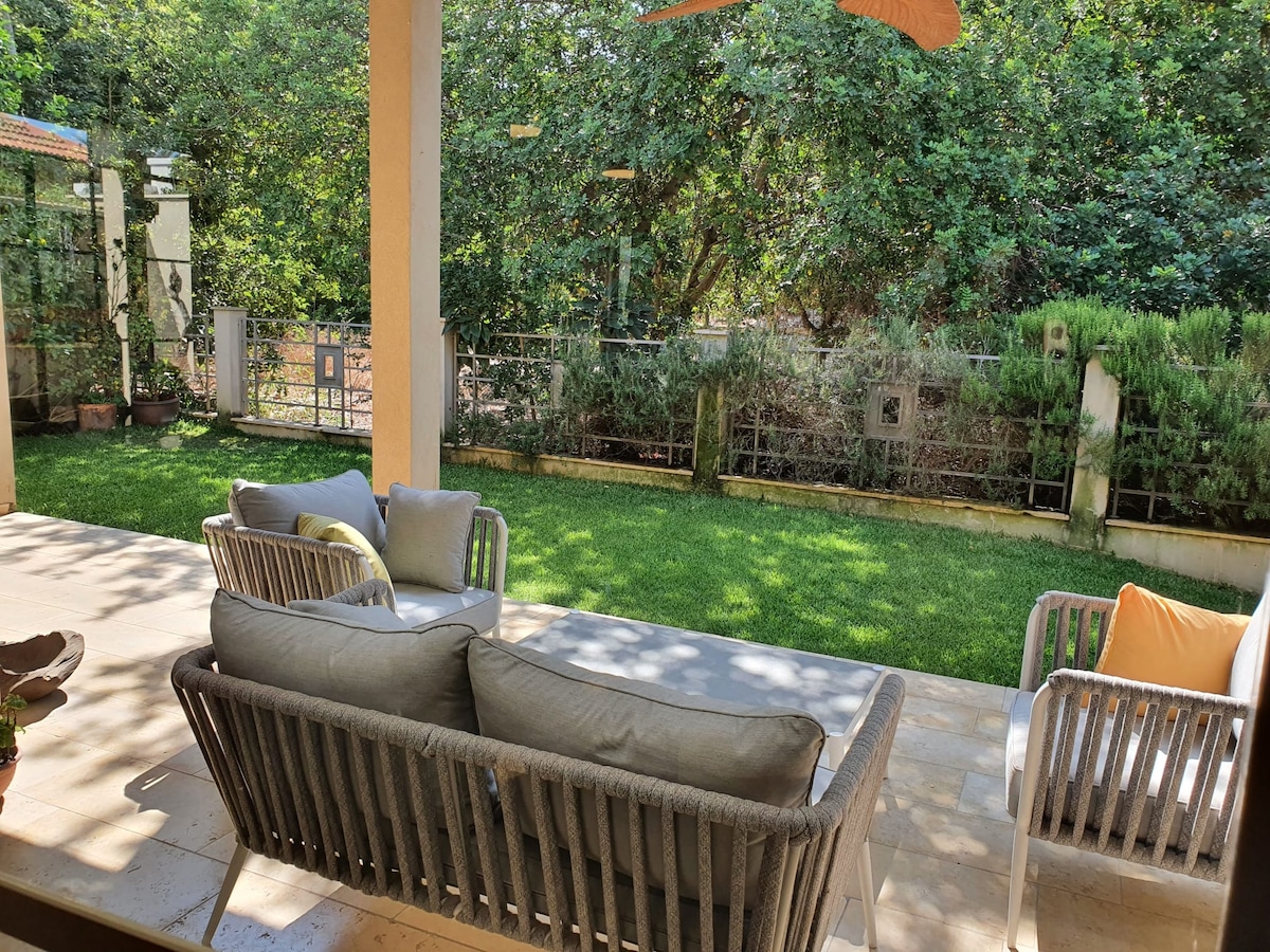 A shaded outdoor seating area is arranged with comfortable, light-colored couches and accent pillows. Lush green grass surrounds the space, complemented by trees and plants in the background, creating a serene environment for relaxation.
