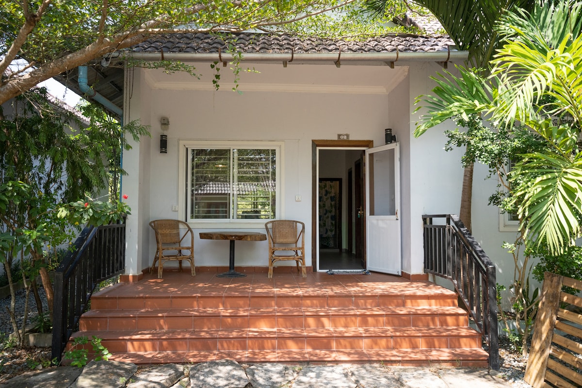 The entrance of the bungalow features a small outdoor patio with a table and two chairs, surrounded by lush greenery. Sunlight filters through the trees, creating a serene atmosphere. The front door is open, inviting guests into the spacious living area.