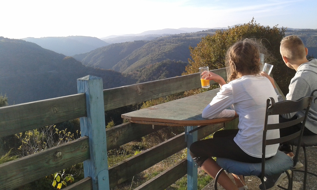 A wooden table with a colorful blue edge is positioned beside a wooden fence. Two individuals enjoy drinks while overlooking a vast valley, with rolling hills and distant mountains visible under a clear sky. The serene landscape evokes a sense of tranquility.