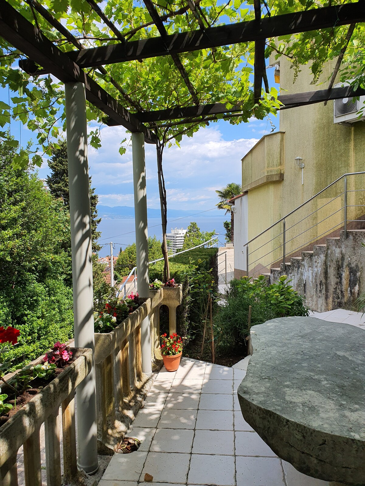 A shaded terrace area is framed by a wooden pergola covered in greenery, providing a serene view of the surrounding landscape. Potted plants and flowers add color to the space, while natural stone elements enhance the outdoor environment.