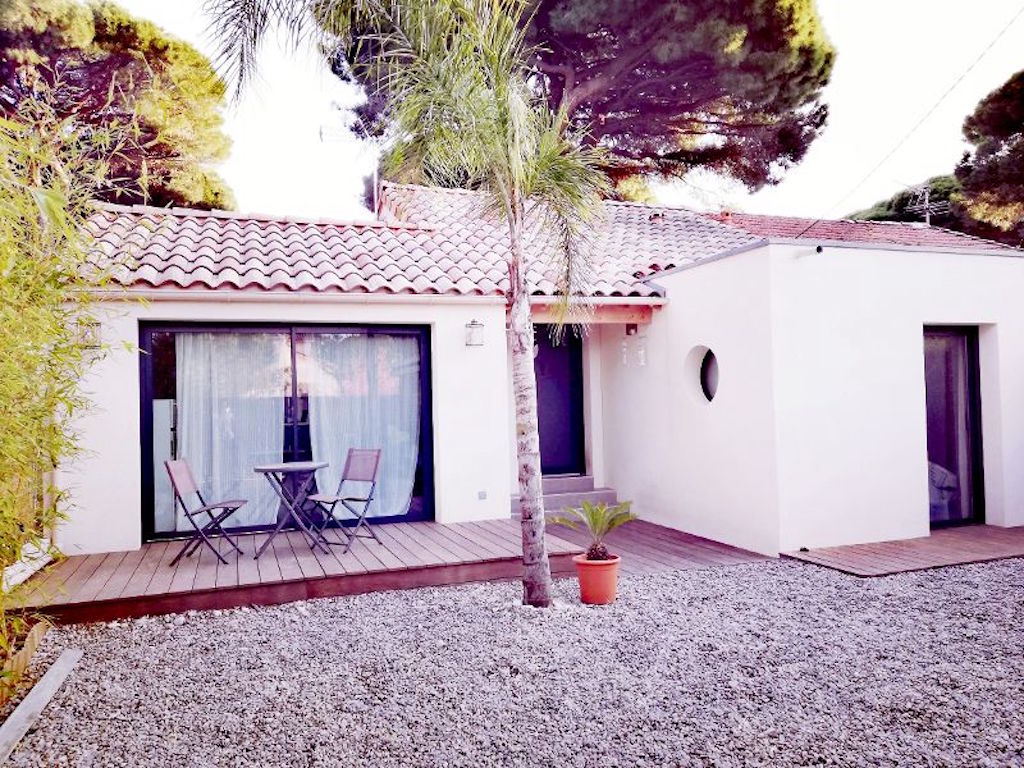 The front of the house features a modern design with a wooden deck and a gray gravel garden. A small round window is visible, along with a palm and potted plant. A patio set comprised of two chairs and a table is positioned on the deck, inviting relaxation.