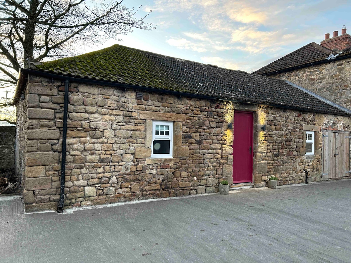 The exterior of a quaint stone building is visible, showcasing rustic charm and a bright red door. Windows are framed in white, providing a contrast against the natural stone facade. A serene sky and tree branches are seen in the background.