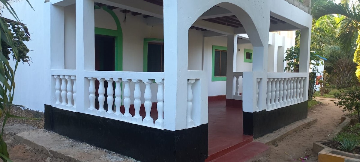 The exterior features a spacious porch with a white railing, highlighting decorative columns. The ground is tiled in red, and the walls are painted white with green accents around the windows. Lush tropical plants are visible nearby, contributing to the serene environment.