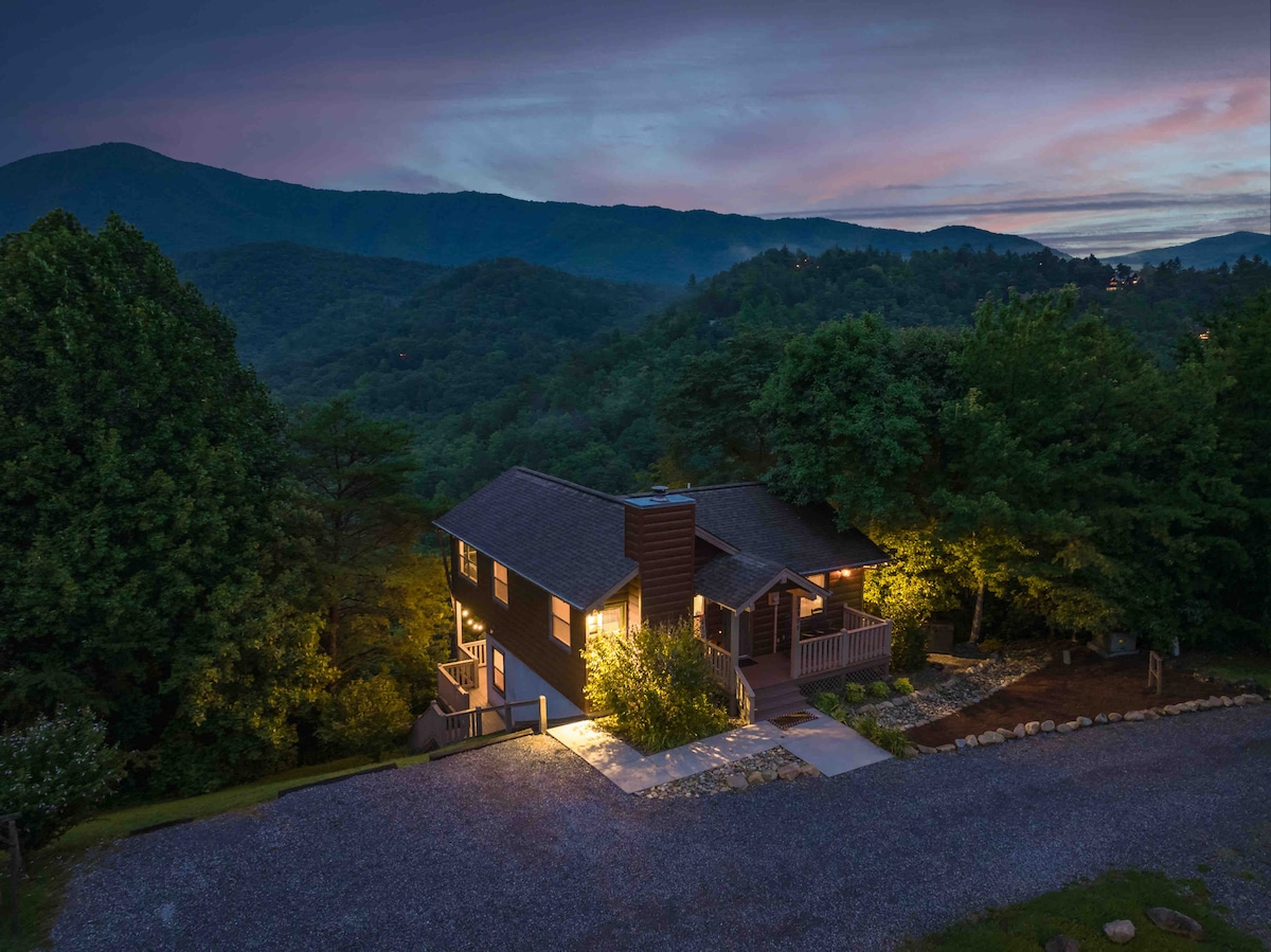 A mountain cabin is illuminated at dusk, nestled among trees. The gravel driveway leads to the entrance, while stunning views of the surrounding hills and valleys are visible in the background, creating a serene connection with nature.