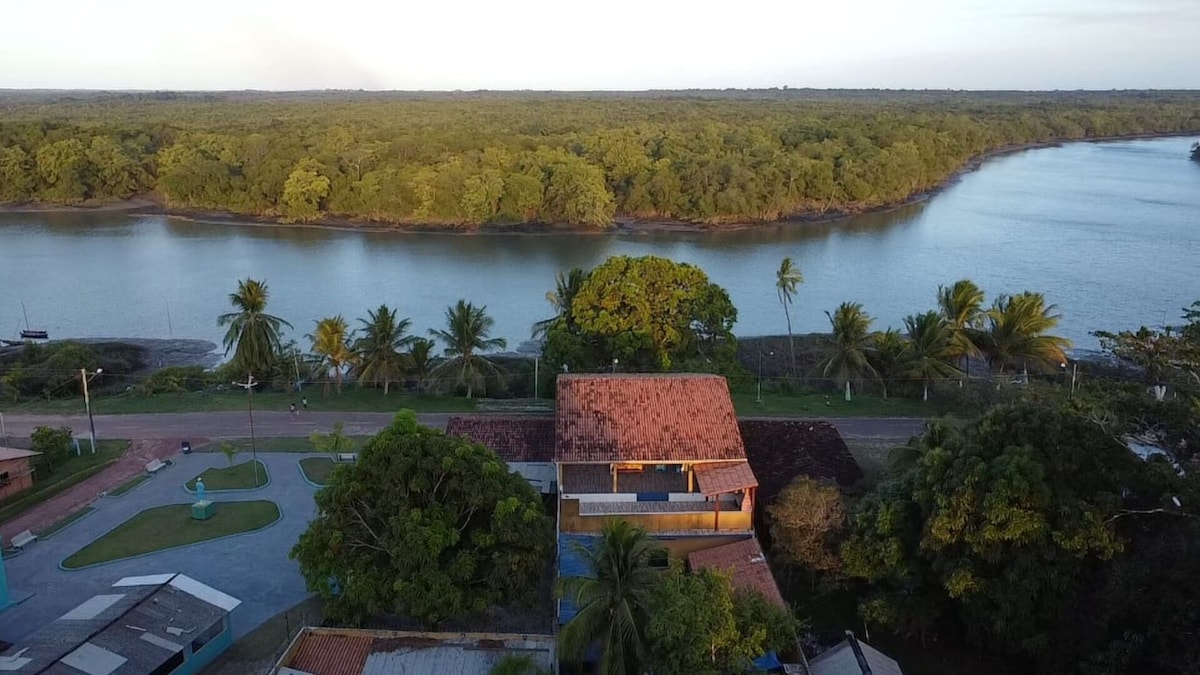 An aerial view of the accommodation reveals a lush green landscape surrounding a winding river. The property features a spacious outdoor area with plants and trees, with the river reflecting soft light from above. The overall setting offers a serene and natural ambiance.