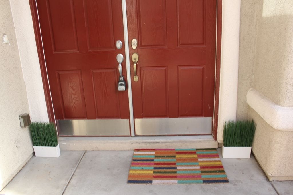 The entrance features a double door with a rich red finish, complemented by a colorful striped doormat. On either side, decorative planters with artificial grass add a touch of greenery to the entryway.
