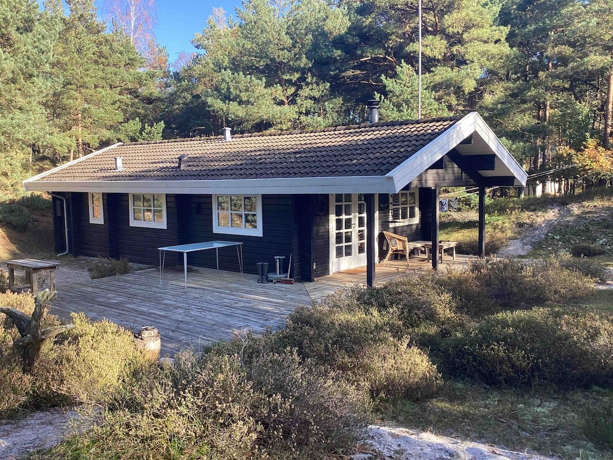 A charming wooden summer house surrounded by a natural landscape. The exterior features large windows and a covered patio area. A wooden table is visible on the patio, along with a bench and outdoor seating. The setting includes trees and low shrubs, emphasizing privacy.