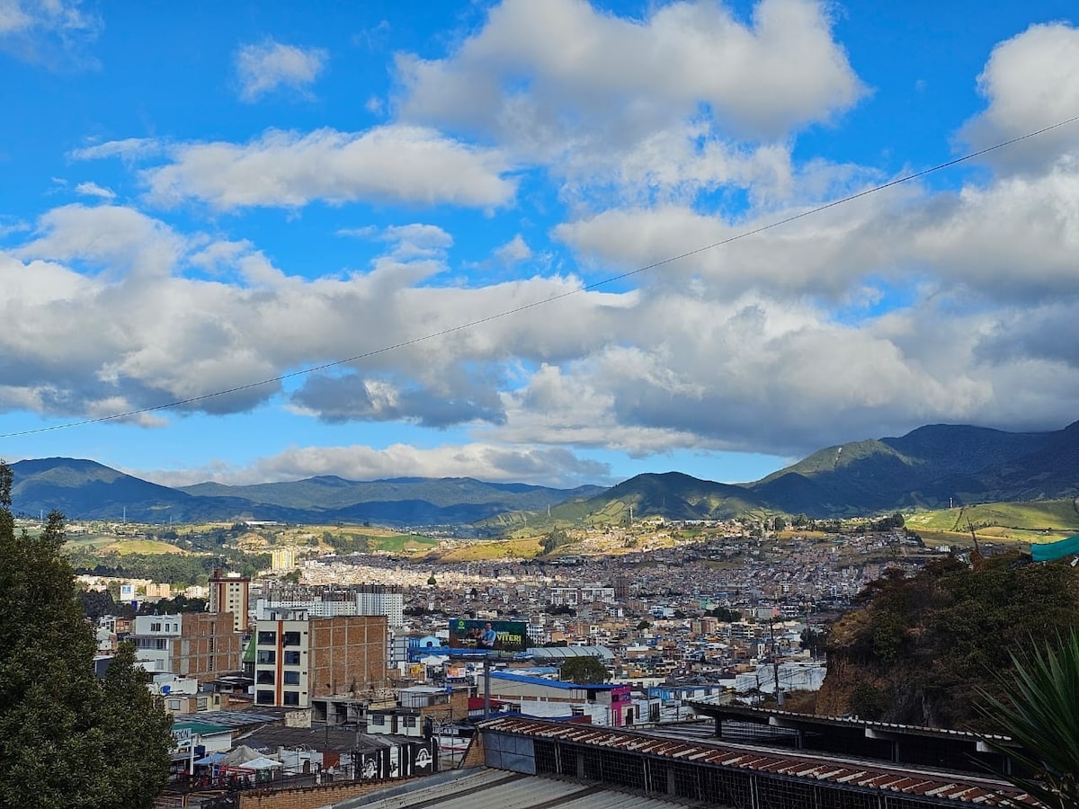 A panoramic view showcases a sprawling city nestled among rolling mountains under a partly cloudy sky. The landscape displays a vibrant mix of urban buildings and natural greenery, allowing for a sense of tranquility within the bustling environment.