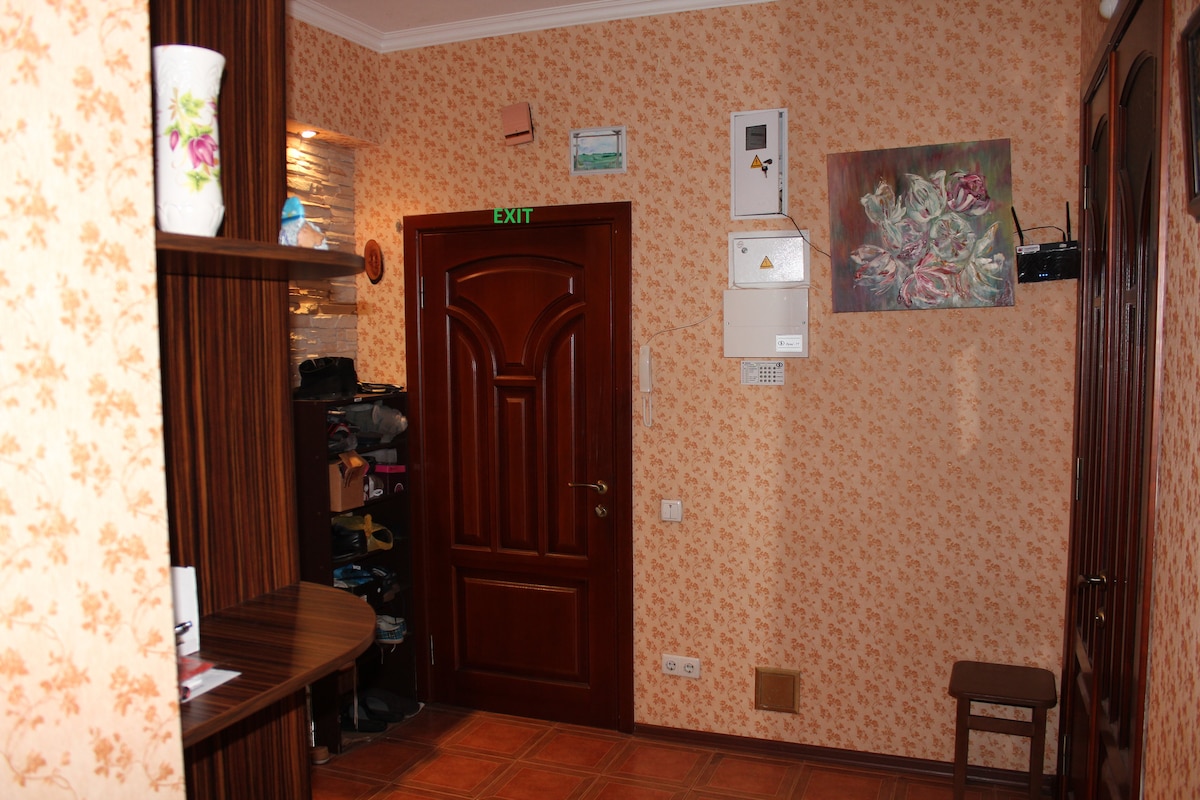 The hallway features floral wallpaper and warm lighting. A wooden shelf is positioned to the left, displaying decorative items. A closed wooden door is visible at the end, while safety and alarm equipment is mounted on the wall. A piece of artwork adds a touch of color.