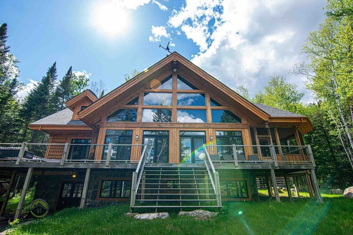The exterior of the chalet features a modern facade with large glass windows framed by wooden beams. A spacious deck with steps leads to the lush green lawn, surrounded by trees. A bright sky with scattered clouds is seen above, enhancing the natural setting.
