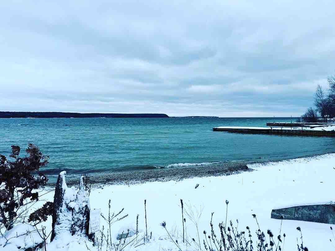 A snowy shoreline is depicted, with calm blue waters meeting a cloudy sky in the background. Pebbles and sand are visible along the edge, leading to the water. The distant shoreline highlights the natural beauty of the area, evoking a serene winter landscape.