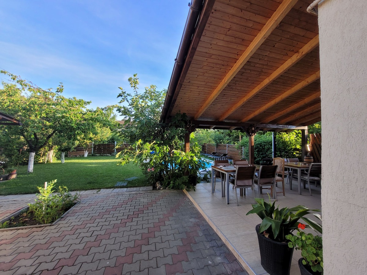 A covered outdoor dining area features wooden beams and ample seating for guests. Lush greenery surrounds the space, with a well-maintained lawn visible in the background. Paved walkways guide towards a swimming pool partially hidden by the foliage, enhancing the serene ambiance.