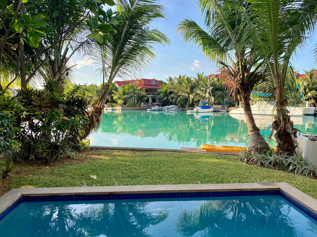 A serene view overlooks the tranquil basin, framed by lush palm trees. The private pool is positioned in the foreground, offering a refreshing contrast against the greenery and the crystal-clear water reflecting the sky. Boat docks can be seen in the distance.